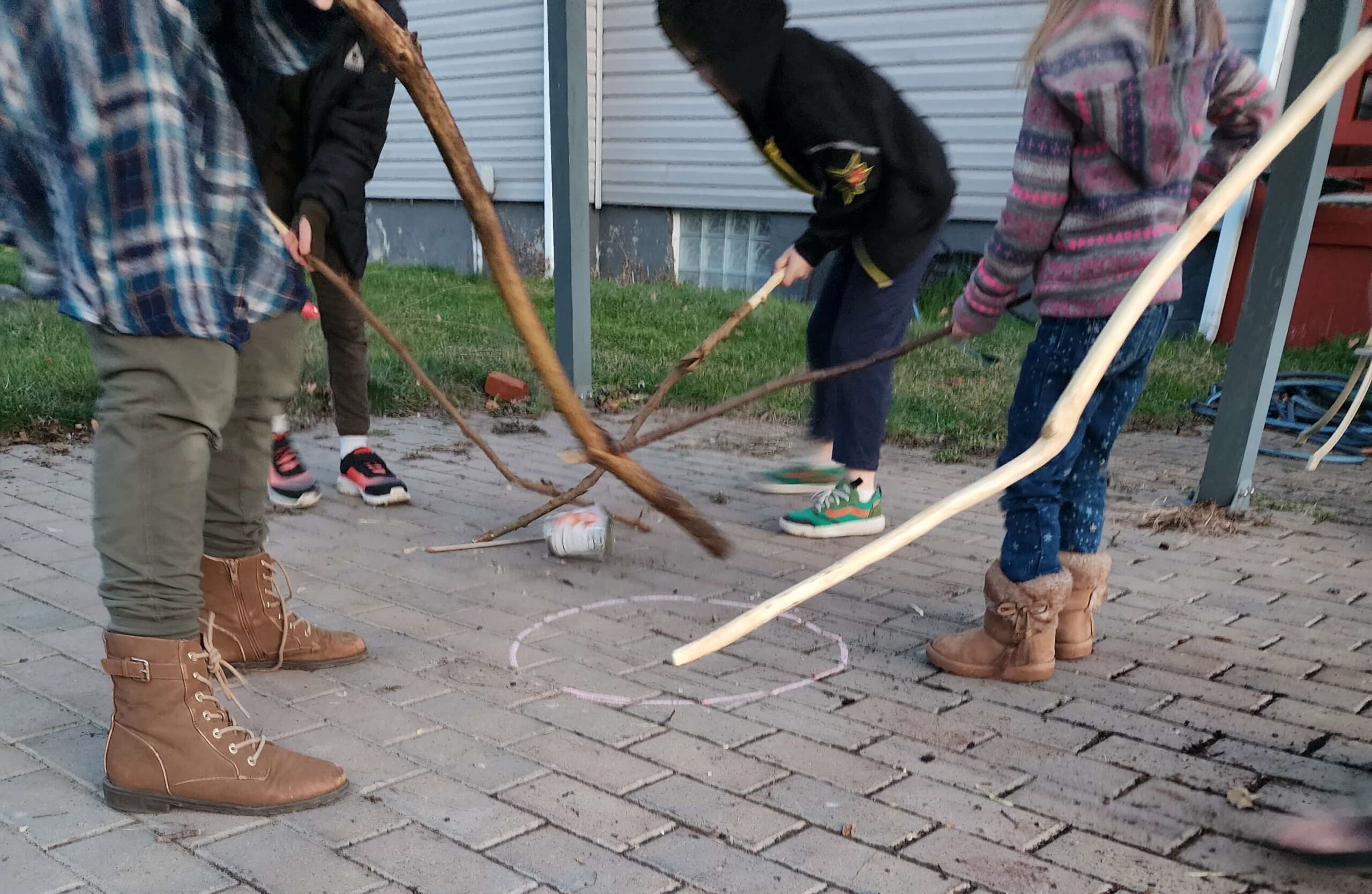 Children swinging sticks at a tin can standing on a cobble stone pavement.