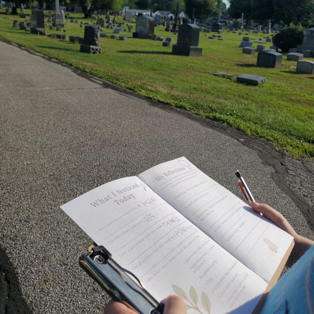 Cemetery journal held in a child's hand with cemetery gravestones amid grass in the background demonstrating cemetery etiquette for families