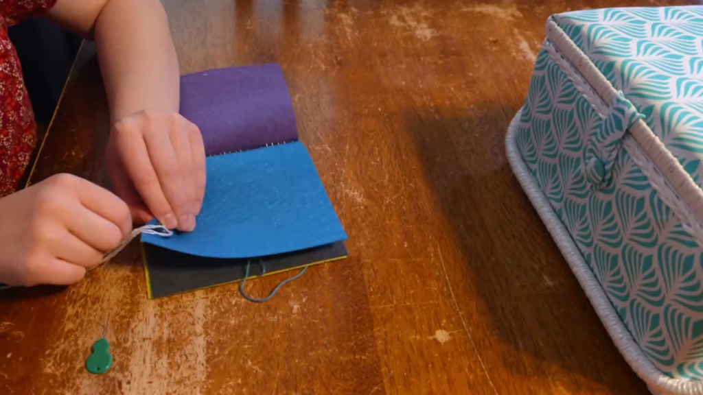Child's hands sewing a button in a felt book as they learn hands-on family history in front of a sewing kit on a wooden table.